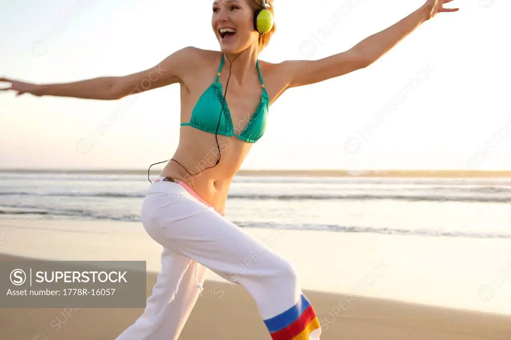 Young female wearing a headset jumps and smiles at the beach.