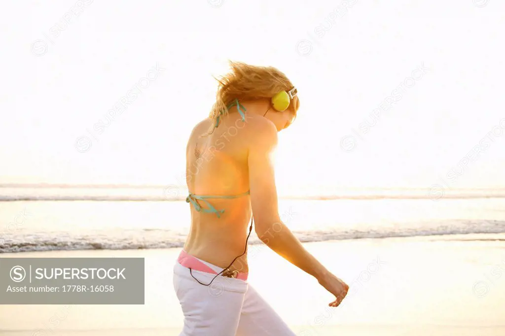 Young female wearing a headset walks away at the beach during sunset.