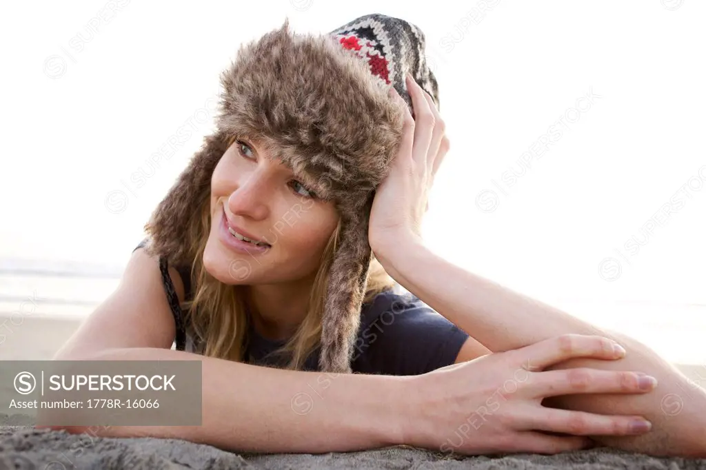 Young female wearing a furry hat lays on sand and supports her head with one hand.