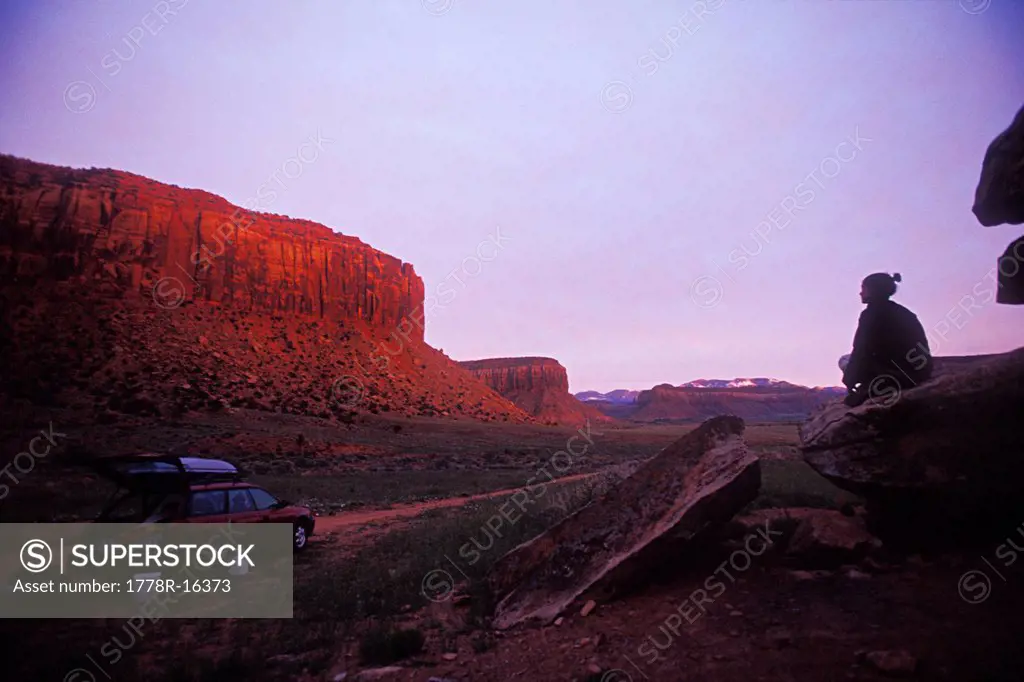 Woman sitting on a rock watches sun set on cliffs above her car.