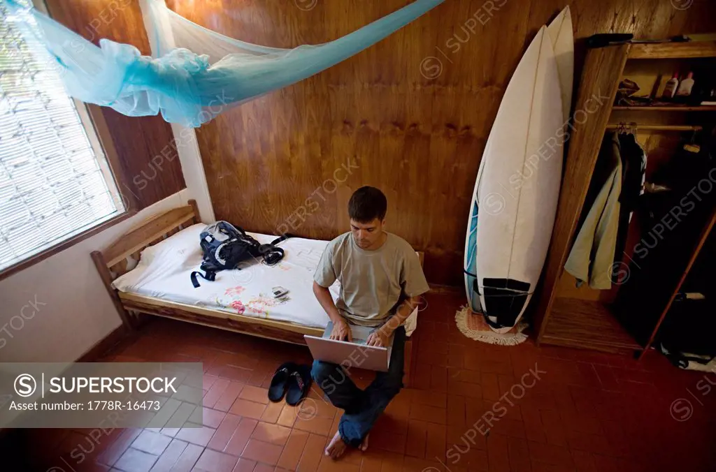 Man sitting on bed works on laptop with surfboards in background.