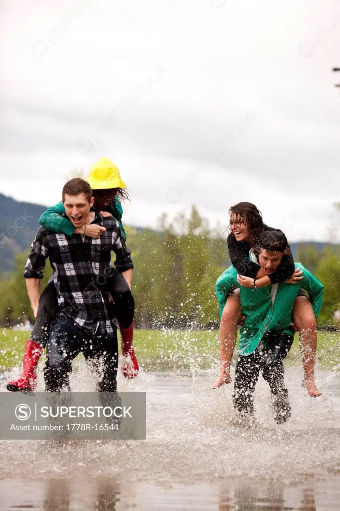 Young adults give each other piggy back rides as they race through a large puddle.