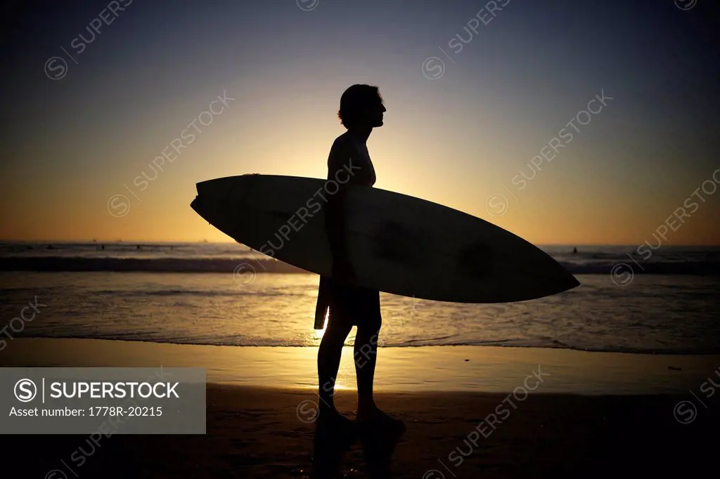 Silhouette of surfer holding a surfboard at the beach during sunset.