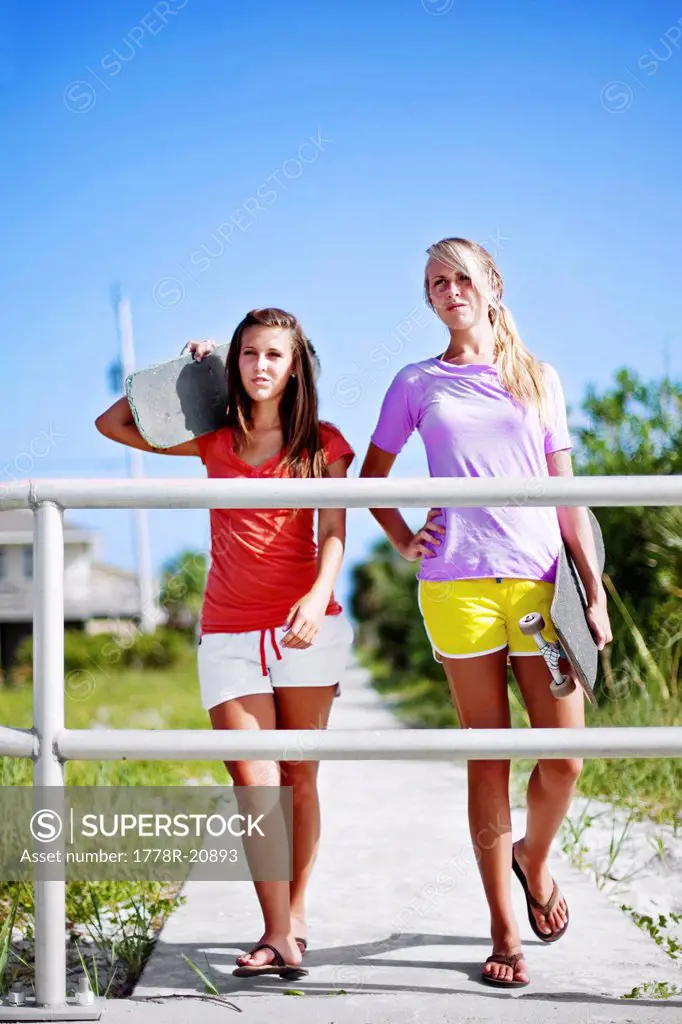 Two girls walk with their skateboards along a sidewalk towards Pensacola Beach, Florida.