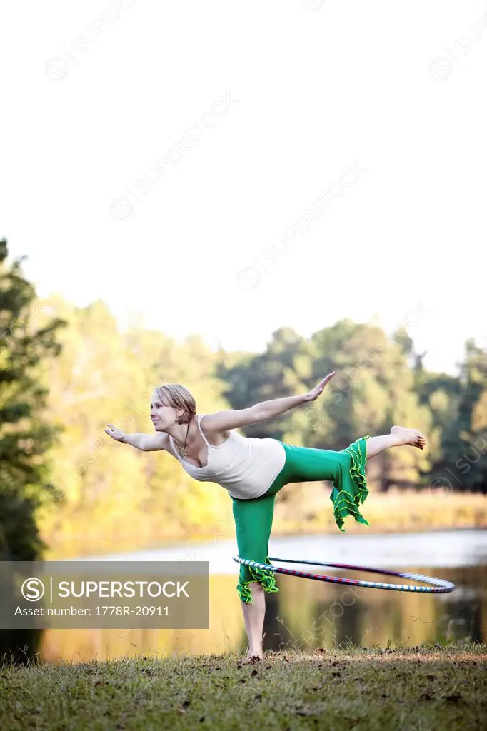 A mid adult woman spins a hoop while standing on one leg with arms out in front of a lake in Chelsea, Alabama. 
