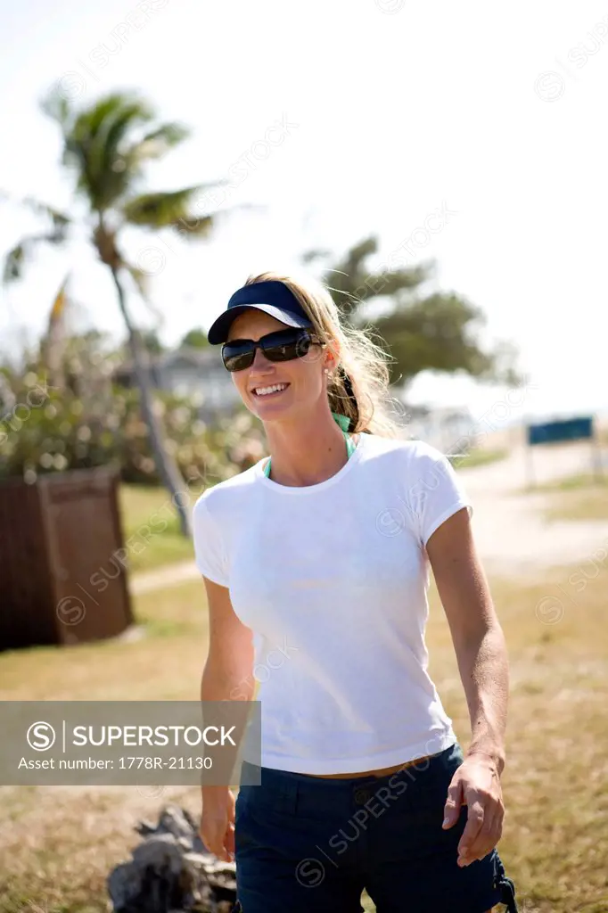 A blonde woman in sunglasses is walking toward the camera with palm trees in the background.