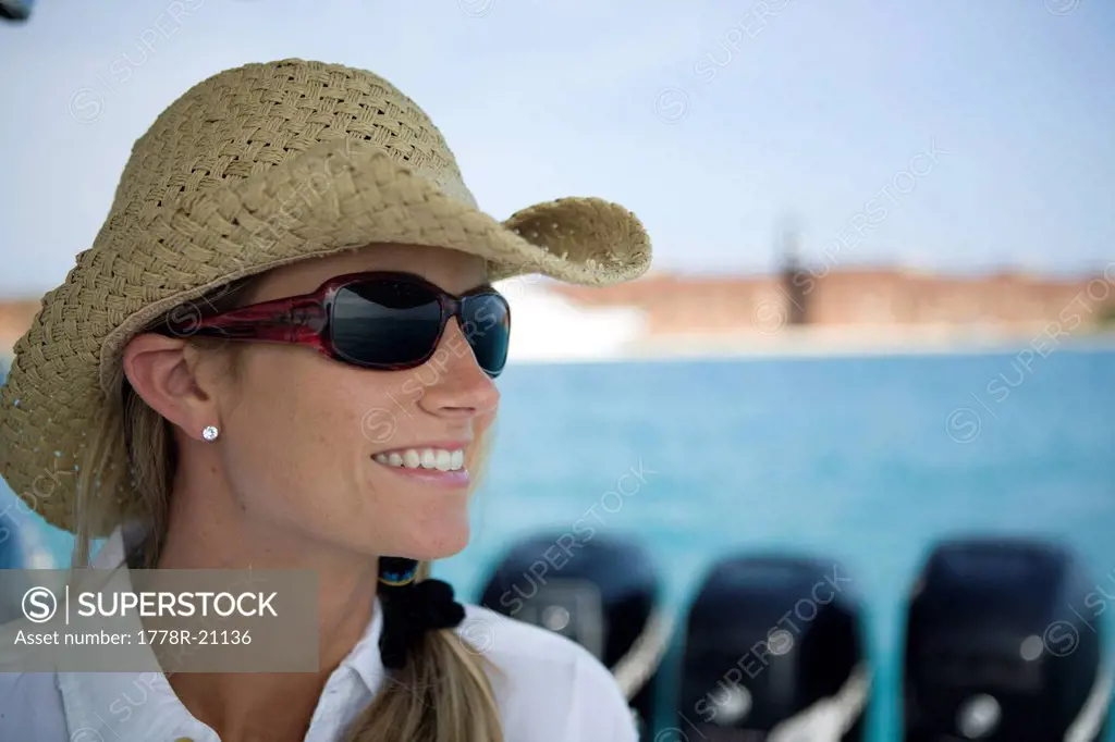 A close_up of a blonde woman as she looks out to the water wearing sunglasses and a straw hat.