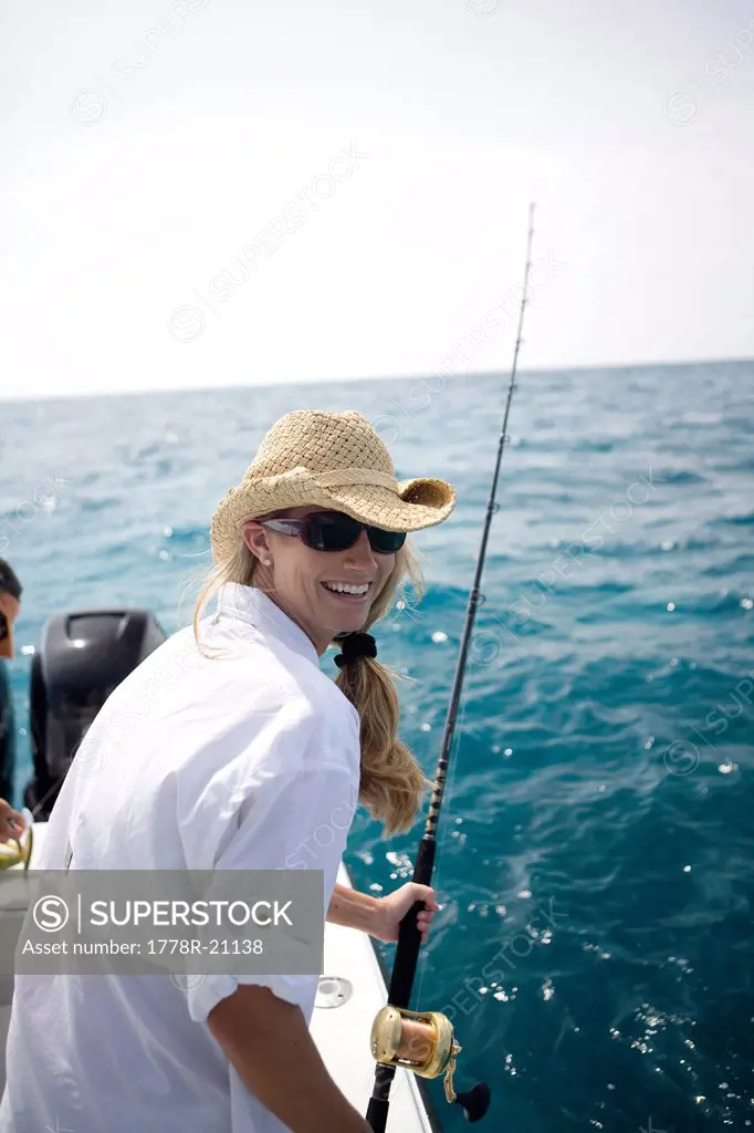 A blonde woman smiles as she holds her fishing rod with blue water in the background.