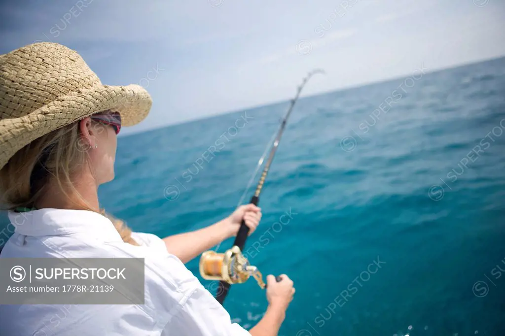 A blonde woman in a straw hat reels in a fish with blue wanter in the distance.