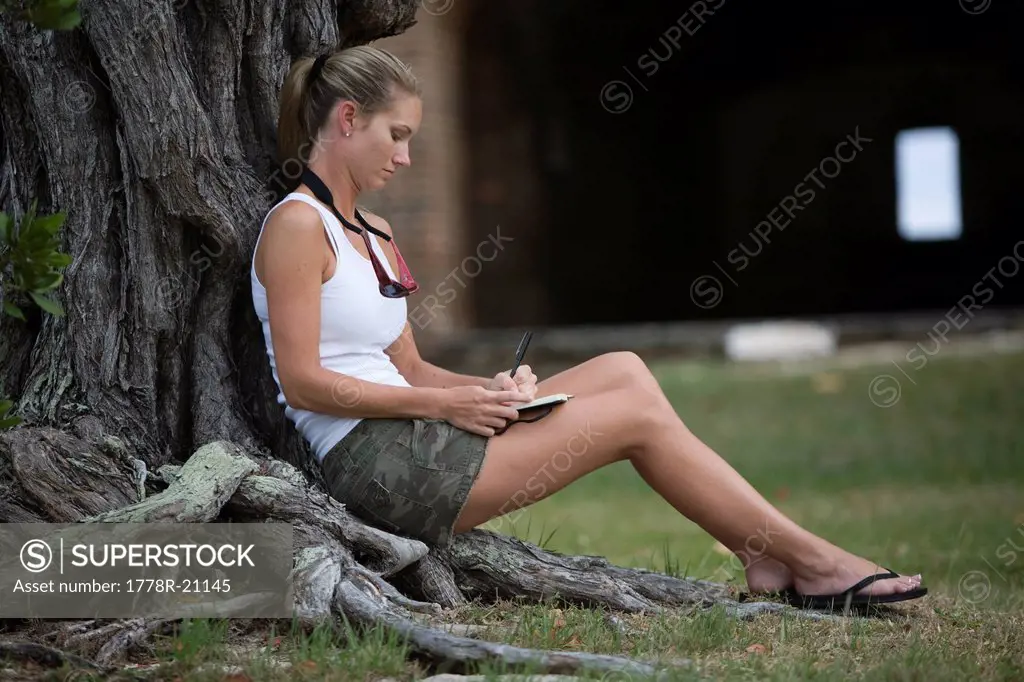 A mid adult woman in shorts is sitting against a tree while she makes a journal entry.