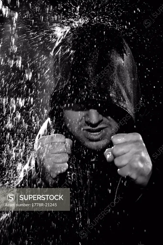 A young man wearing a hooded jacket poses in a wushu stance while it rains down on him.