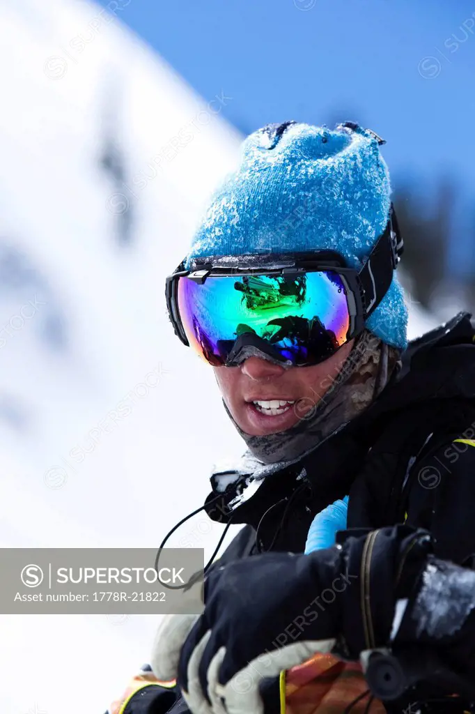 A portrait of a snowboarder smiling with a reflection of a snowmobile in his goggles in the backcountry of Colorado.
