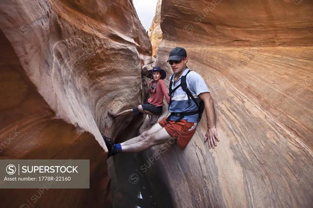 A couple stemming between two walls gazes down canyon while descending a slot canyon in Utah.
