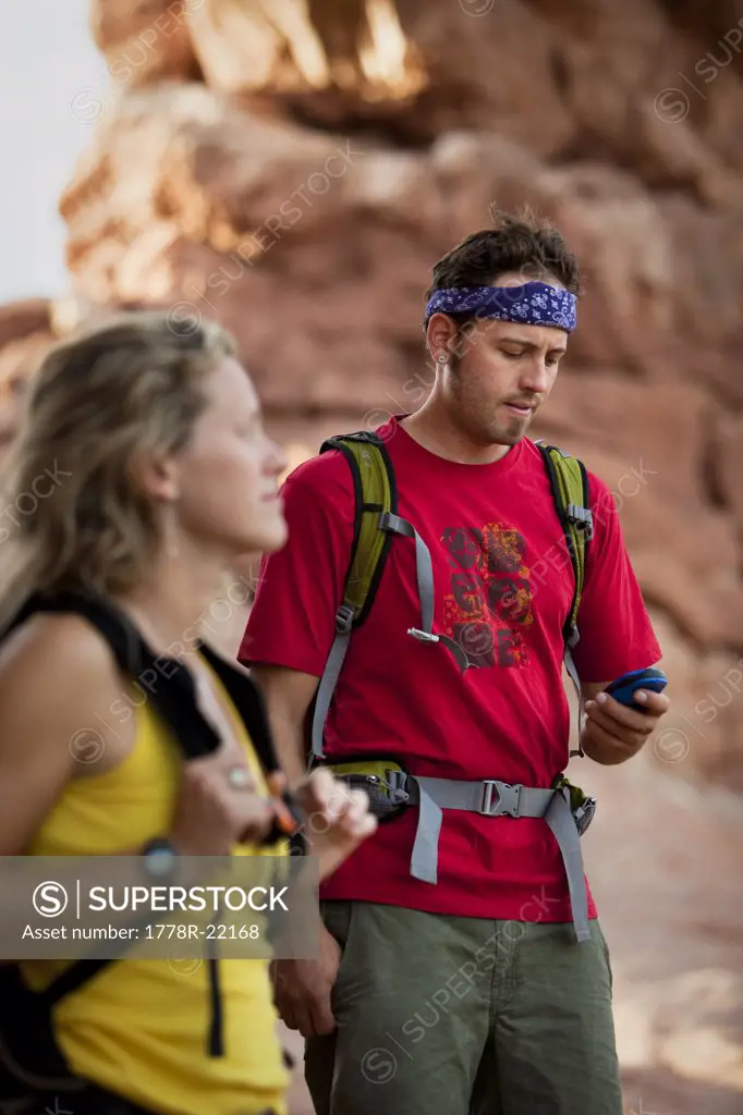 A man with backpack on looks at his GPS while an out of focus woman in the foreground looks on.