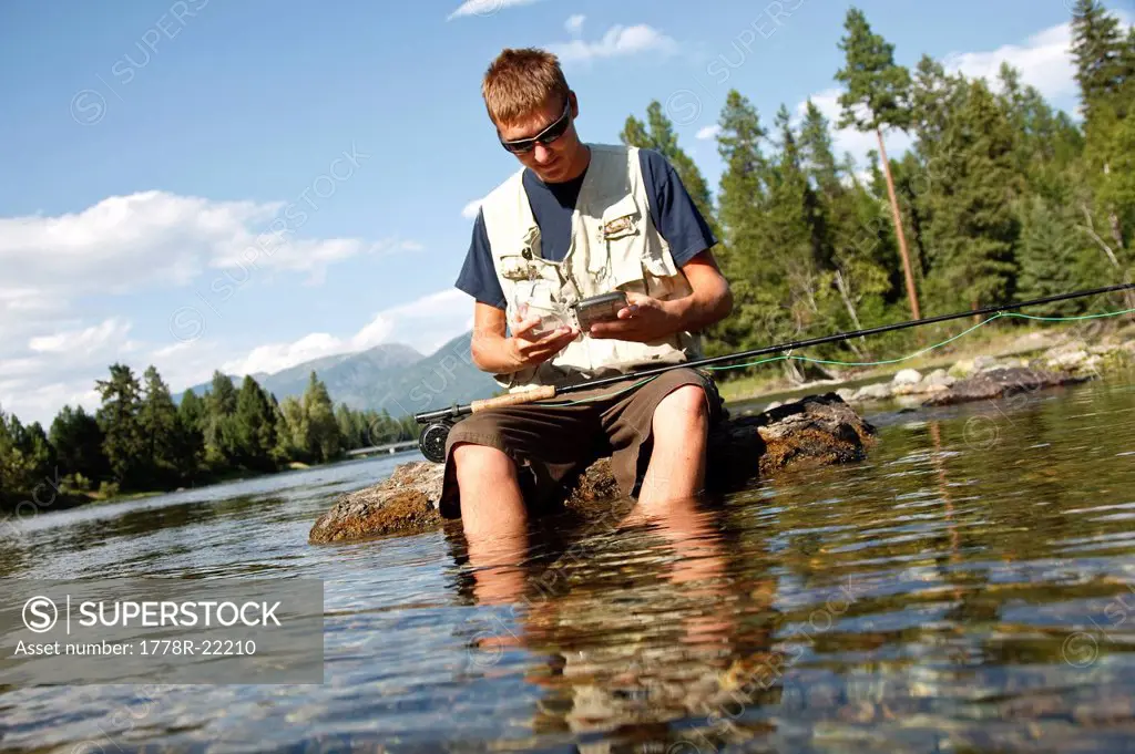 A teenage boy sits on a rock as he chooses a fly while fly fishing on the Swan River near Bigfork, Montana.
