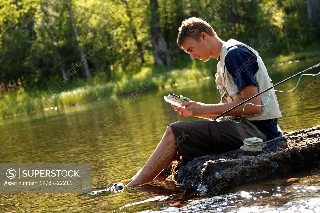 A teenage boy sits on a rock as he chooses a fly while fly fishing on the Swan River near Bigfork, Montana.