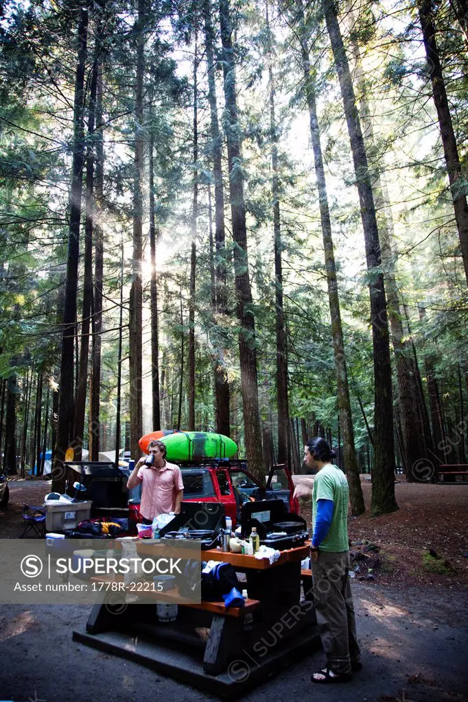 Two men car camping as the sun rays filter through the trees over their campsite.