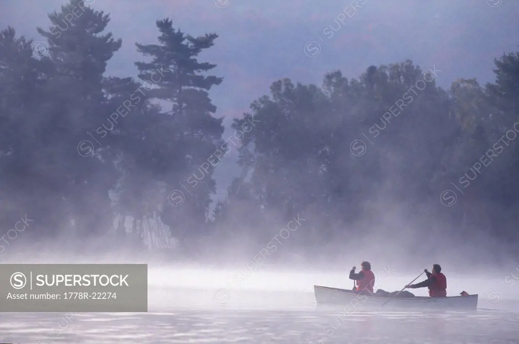 Young/middle_aged couple paddling canoe on misty lake at sunrise near Algonquin Park, Muskoka, Ontario, Canada.