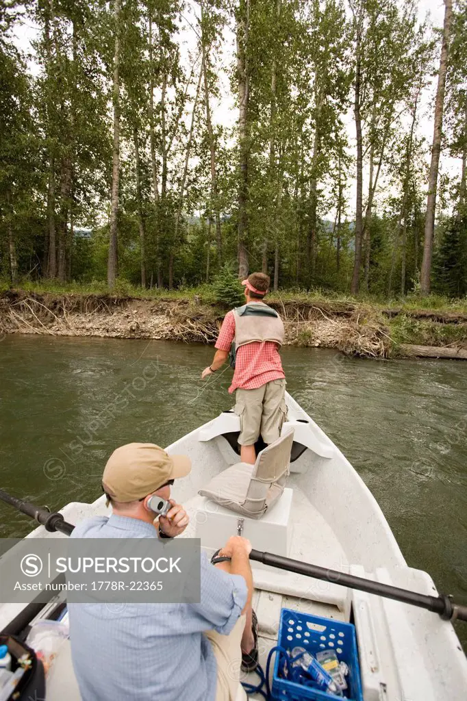 Young man using cell phone while fly fishing on the Elk River from a dory, Fernie, East Kootenays, British Columbia, Canada.