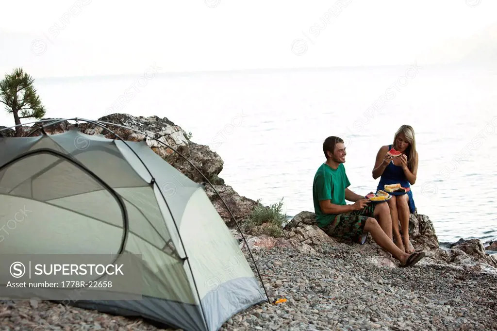 Two young adults camping smile while eating dinner next to a lake in Idaho.