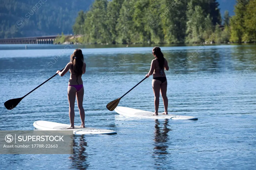 Two athletic young women stand up paddle board on a lake in Idaho on a sunny day.