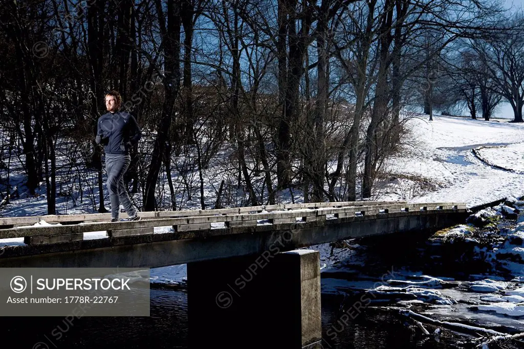 A man runs on a bridge over a creek in the snow.