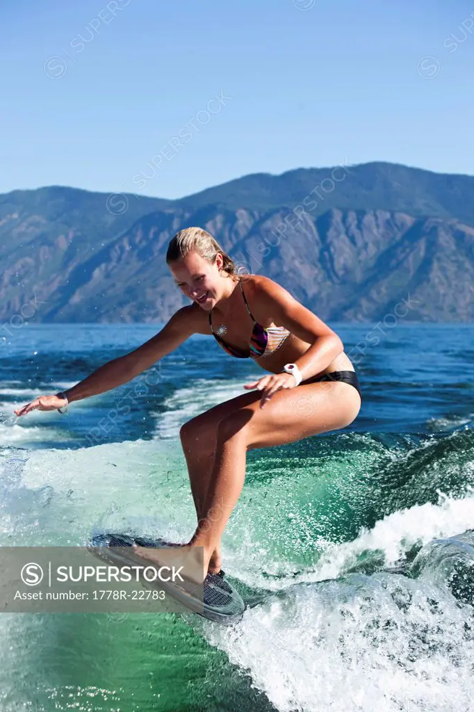 A athletic young woman smiles while wakesurfing behind a wakeboard boat on a sunny day in Idaho.