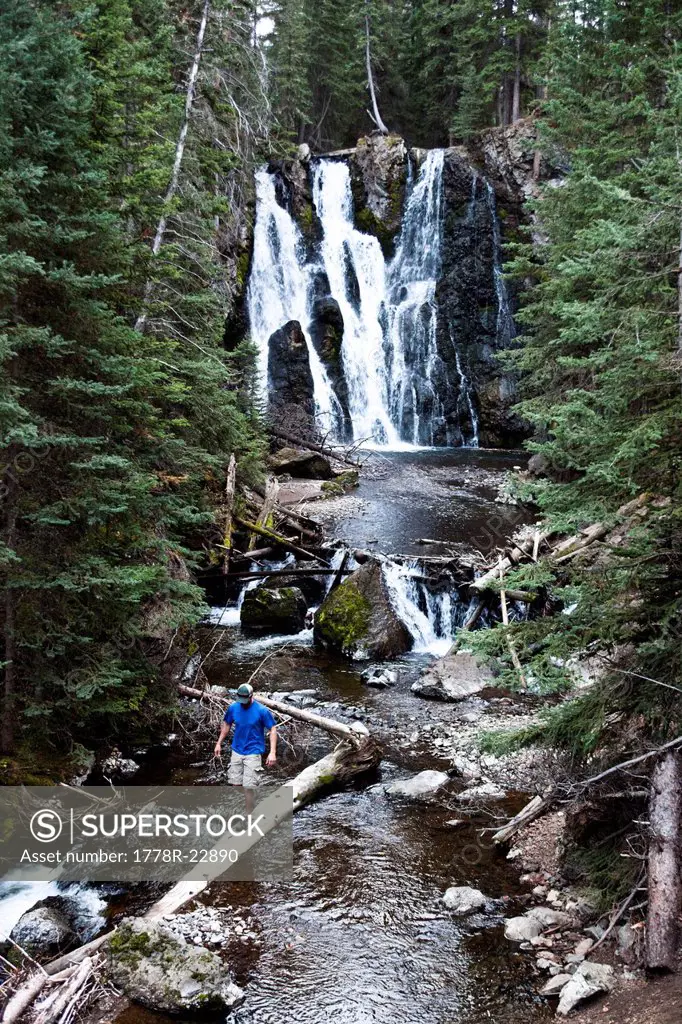 A athletic man hiking walks across a fallen log next to a waterfall in Montana.
