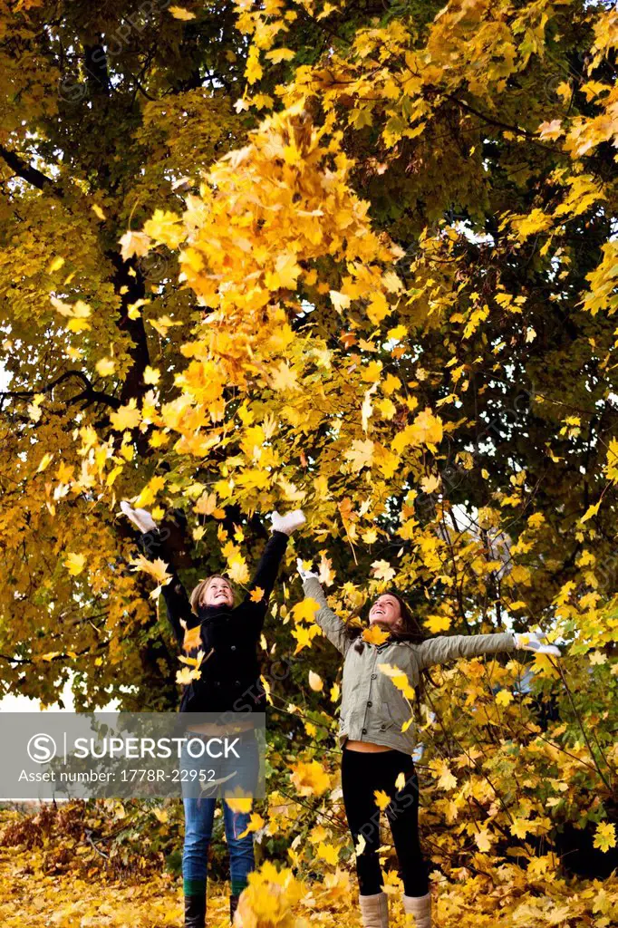 Two young women smiling throw orange leaves into the sky surrounded by fall colors in Idaho.