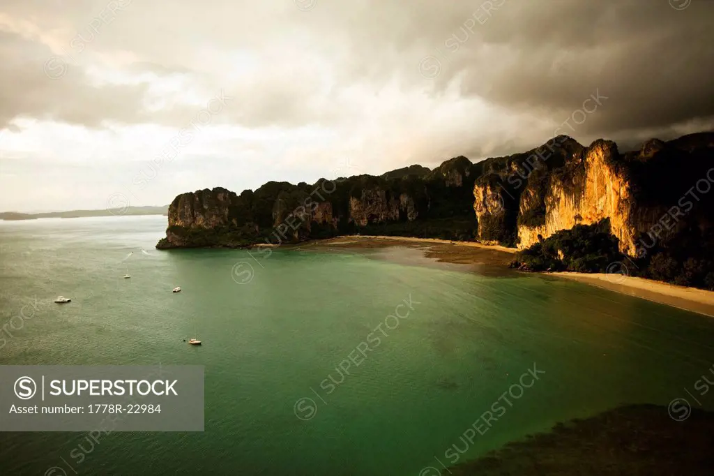 A aerial view of two large white sandy beaches at sunset surrounded by limestone cliffs and turquoise water filled with longtail boats in Thailand.