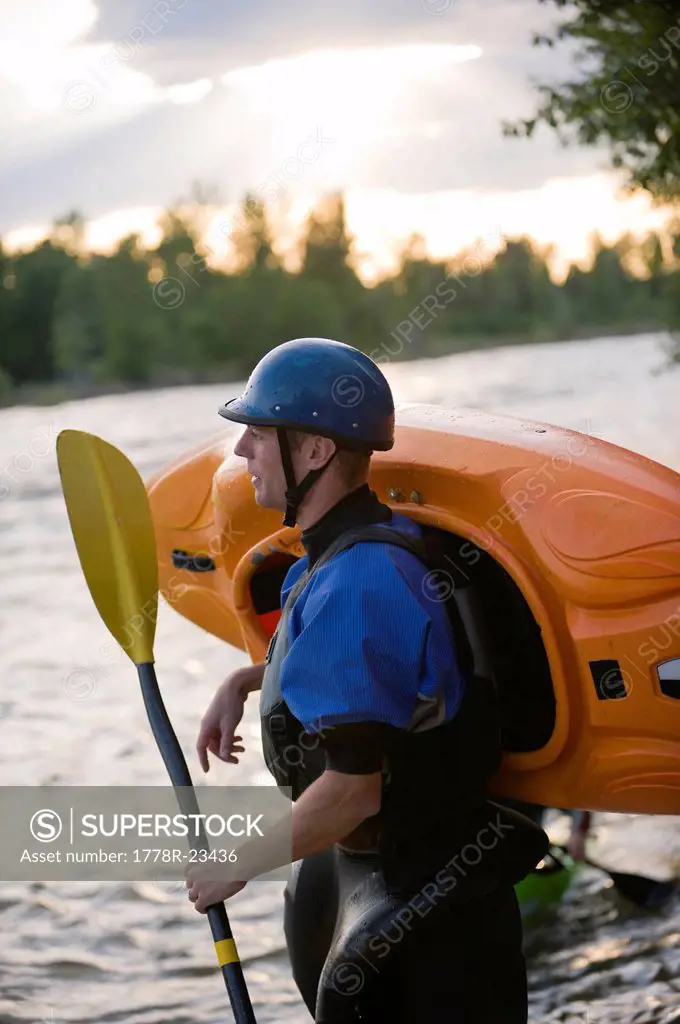A young male kayaker stands along the shore of a river.