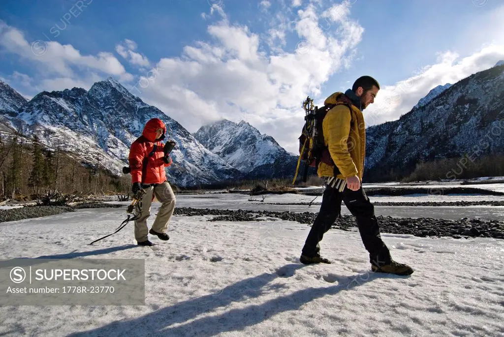 Two young ice climbers walk across a snowy river, with gear in hand, on their way to an afternoon ice climb in Echo Bend, Alaska.