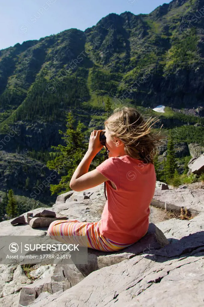 A young girl sits on a rocky outcrop and looks through her binoculars in Glacier National Park, Montana.