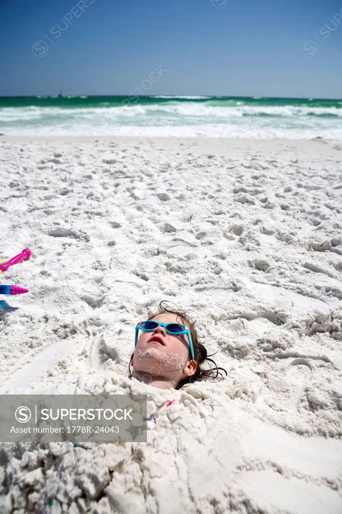 A little girl is buried in the sand up to her head at the beach with the ocean in the background.