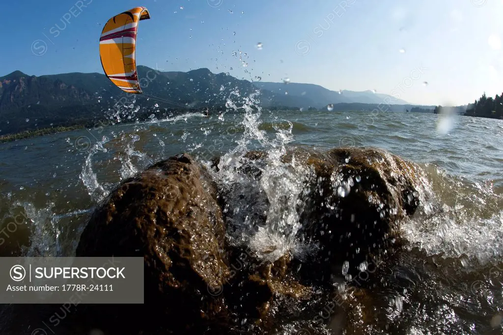 Low angle perspective of a kiteboarder riding by with waves splashing on a rock prominent in the foreground.