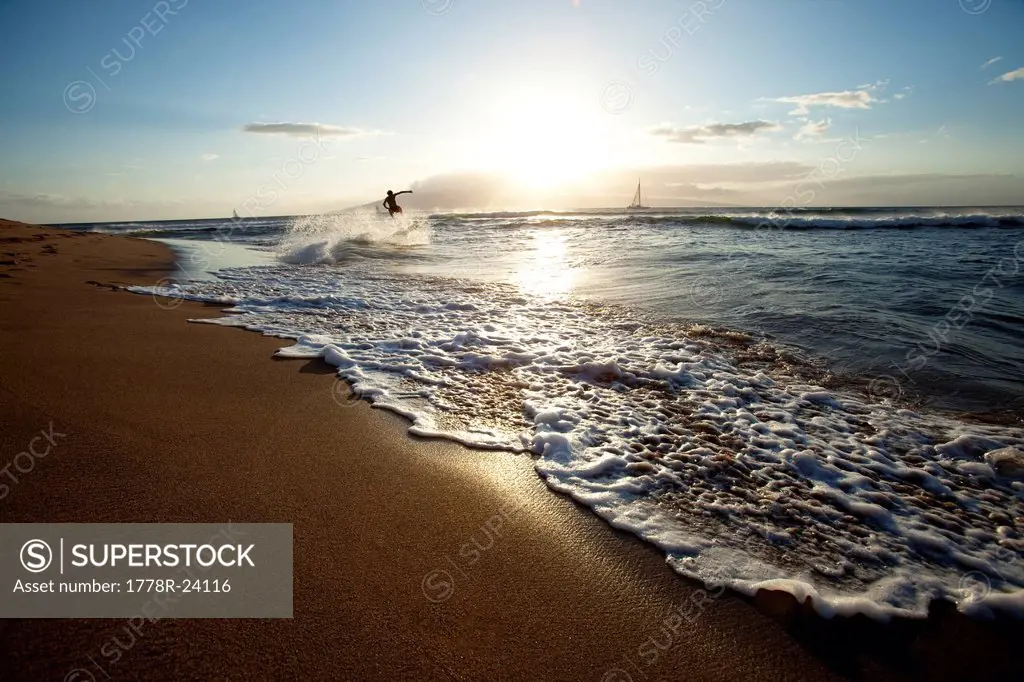 One man skimboarding at sunset with sailboats in the background.