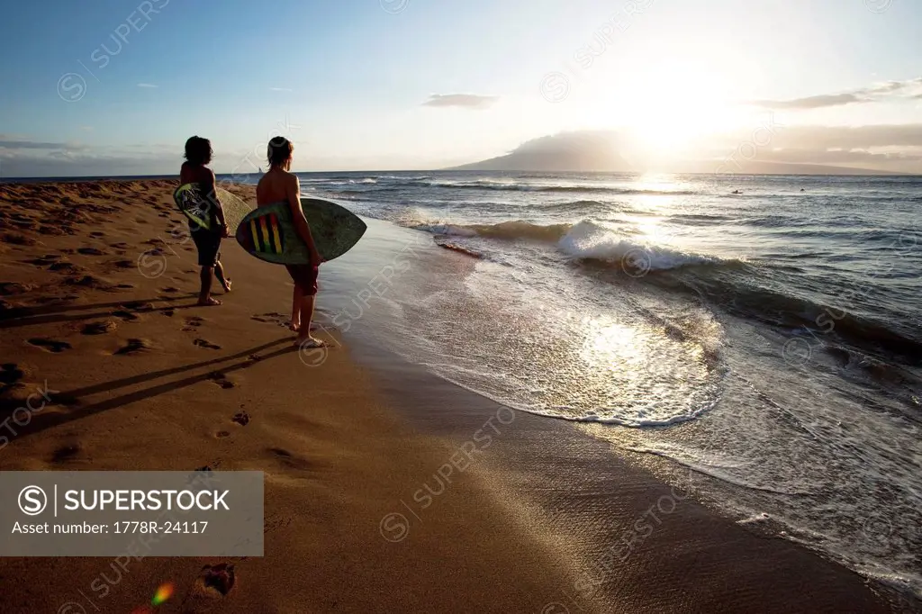 Two men watching the surf at sunset with their skimboards in their arms.