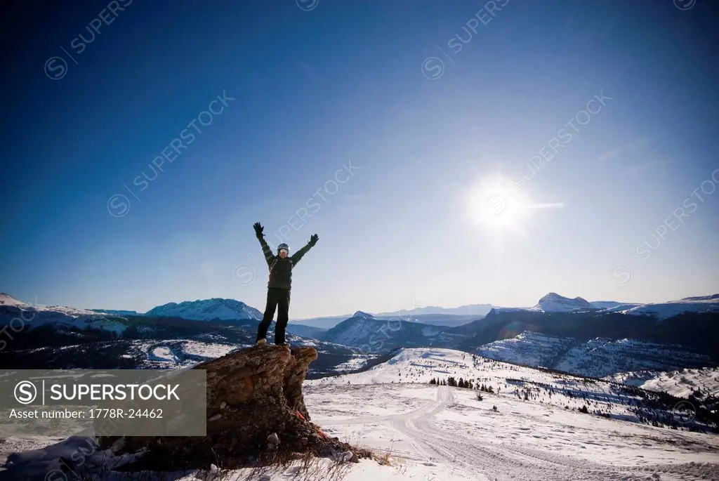 A middle aged man celebrates reaching a high point overlooking a snowy valley in the Rocky Mountains.