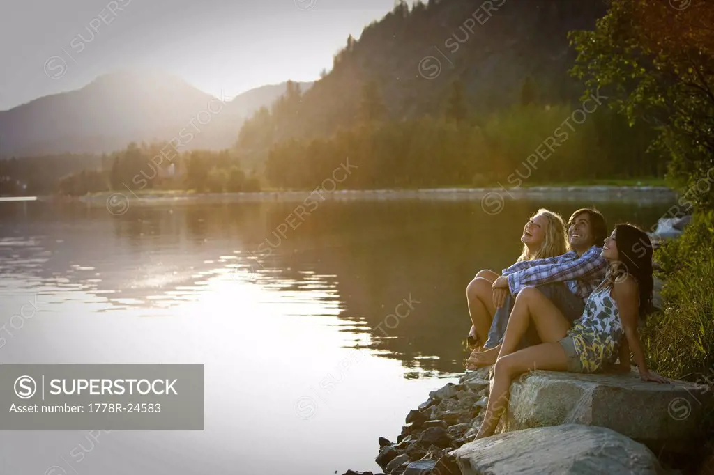Three young adults laugh while sitting on rocks watching the sunset over the lake.
