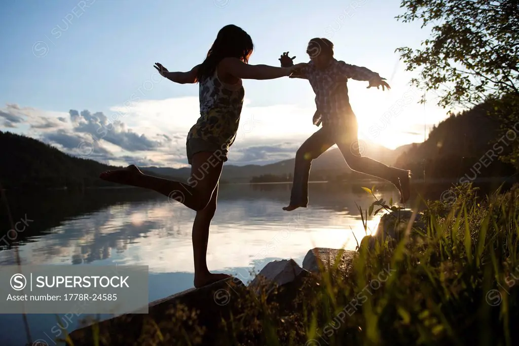 Two young adults balancing on rocks at sunset next to the lake.