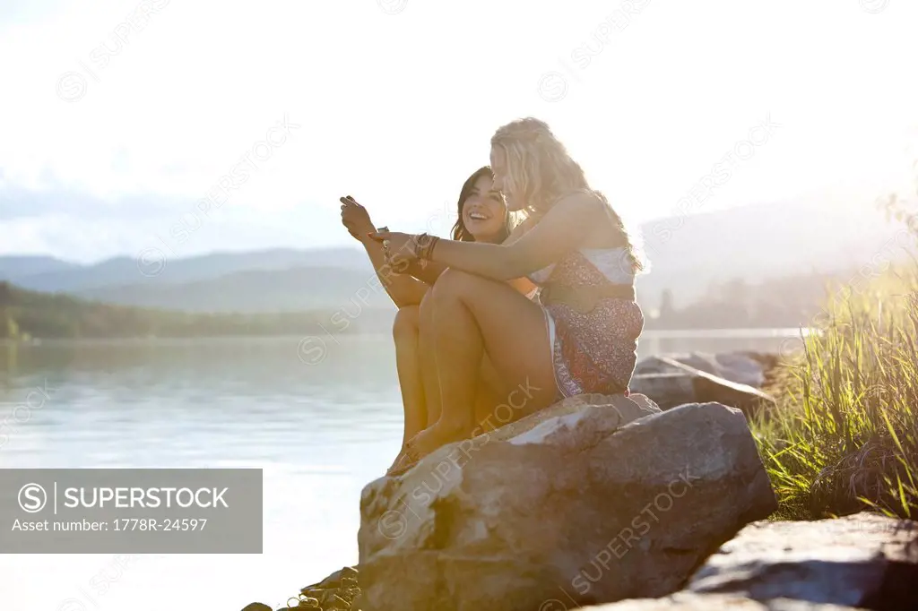 Two young women laugh while texting at sunset on the lake.