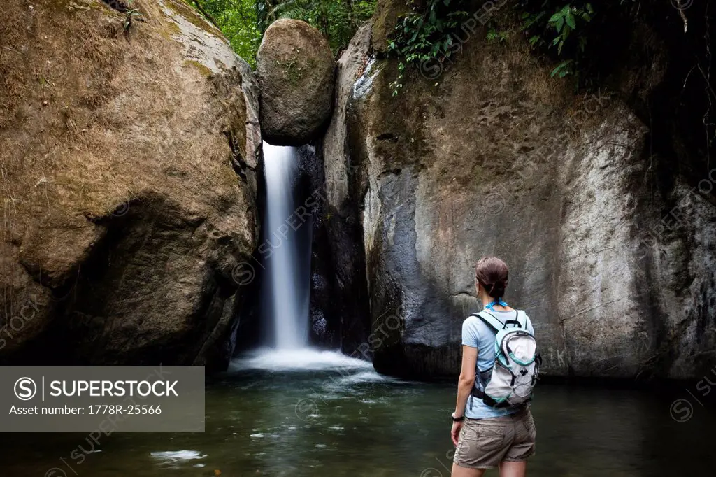A woman stands at the edge of a pool of water looking at a waterfall while wearing a backpack.