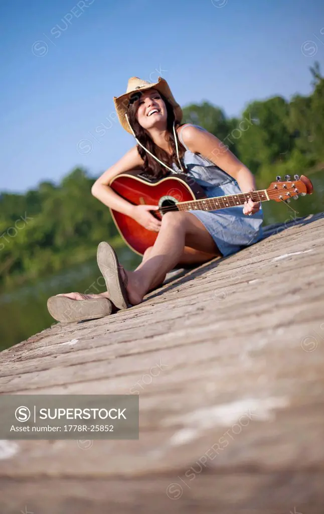 A young woman sits on a pier playing guitar.