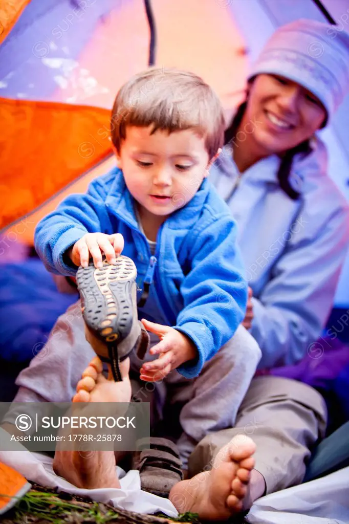 a mother and son sit in a blue and orange tent with their bare feet sticking out the door. the boy tries to put his shoes on. Their campsite is at app...