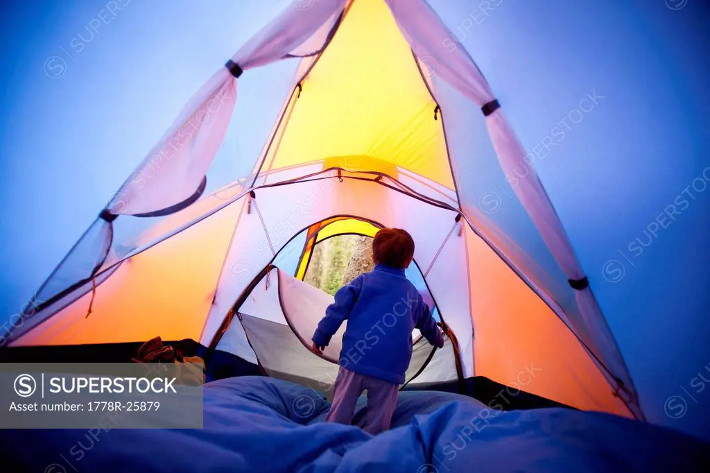 a little boy opens and closes the zippered tent door while he stand on a blue sleeping bag. The tent is orange and blue. The campsite is at approximat...