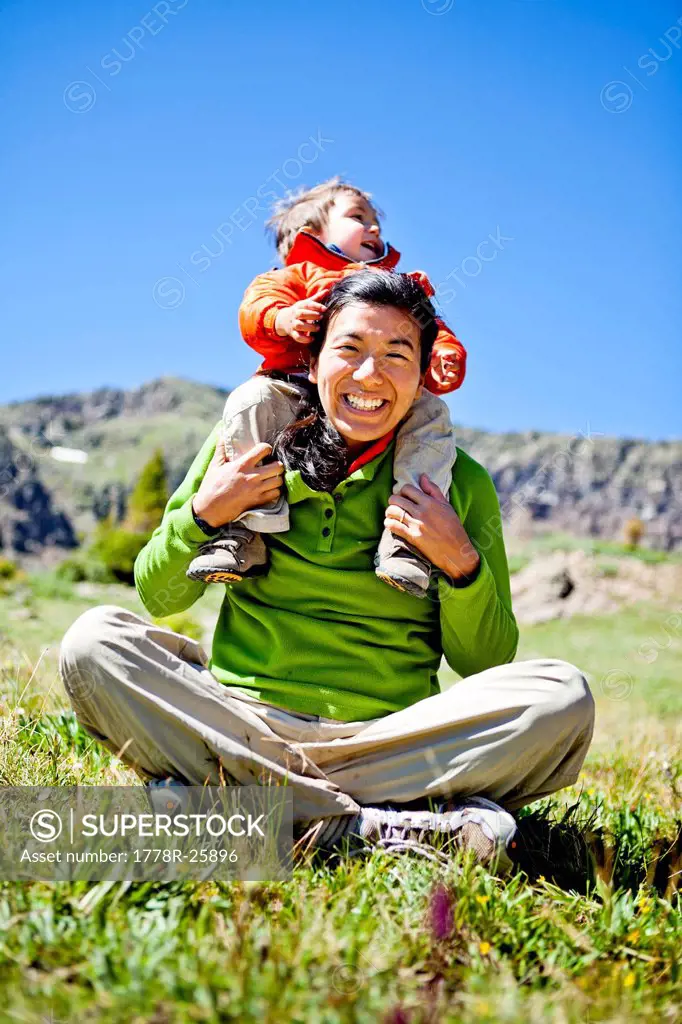 a woman smiles as she sits cross legged in an alpine meadow with wildflowers doing a Buddha pose. Her son, a 2 year old, crawls on her. They are just ...