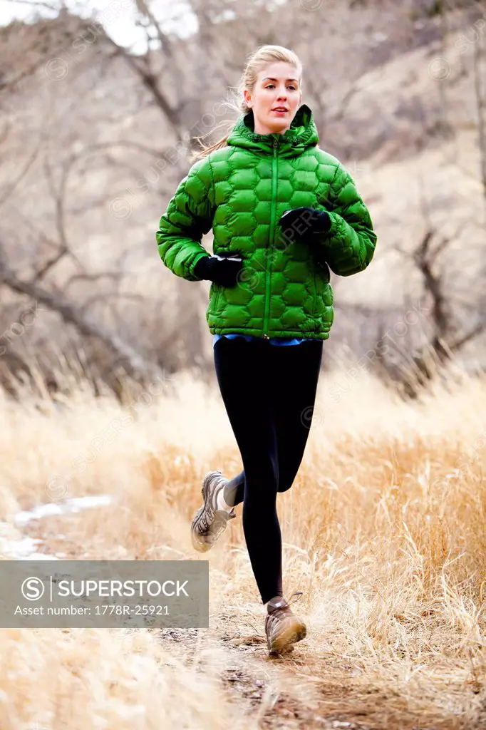 A young woman runs a down trail through dead grass just north of Dixon Reservoir, on a cold day in a green down jacket.