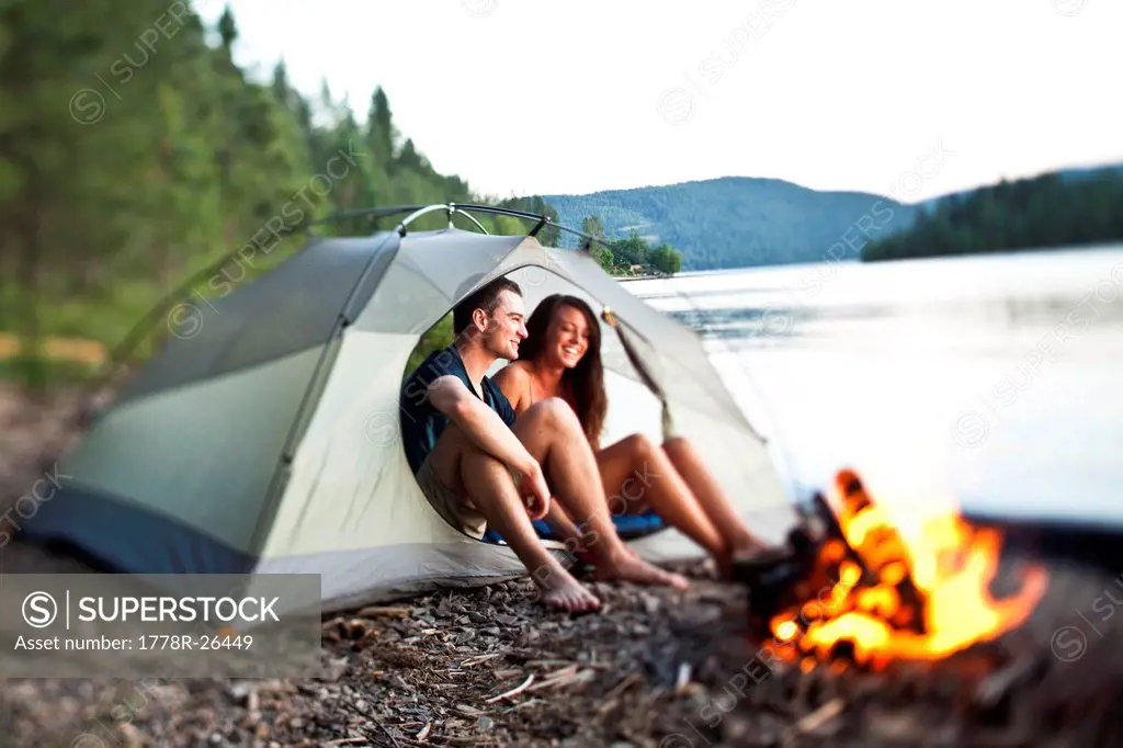 A young adult couple laugh and smile sitting in their tent next to a campfire in Idaho.