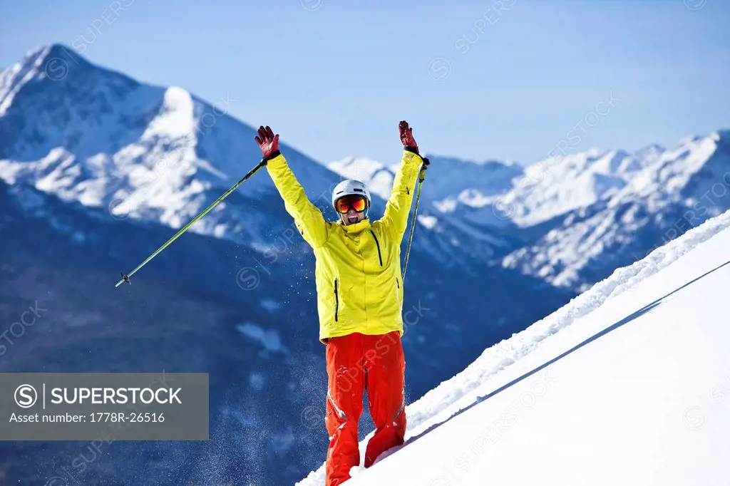 A happy skier smiling raises his arms in celebration of fresh snow with huge mountains behind him in Colorado. 