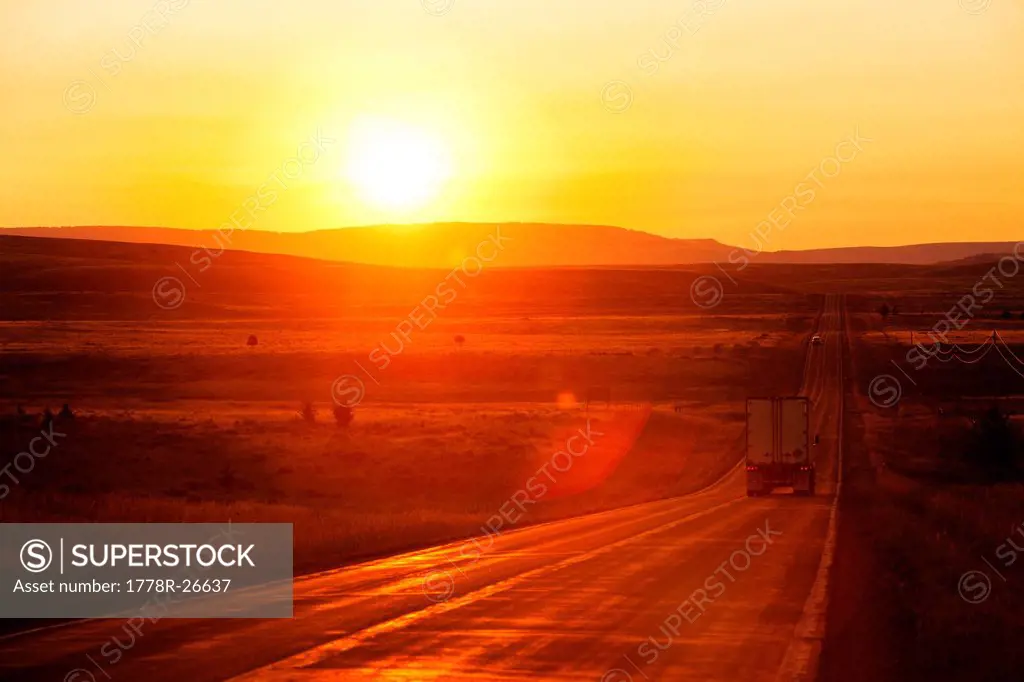 A shipping truck on a long straight highway at sunrise in Montana.